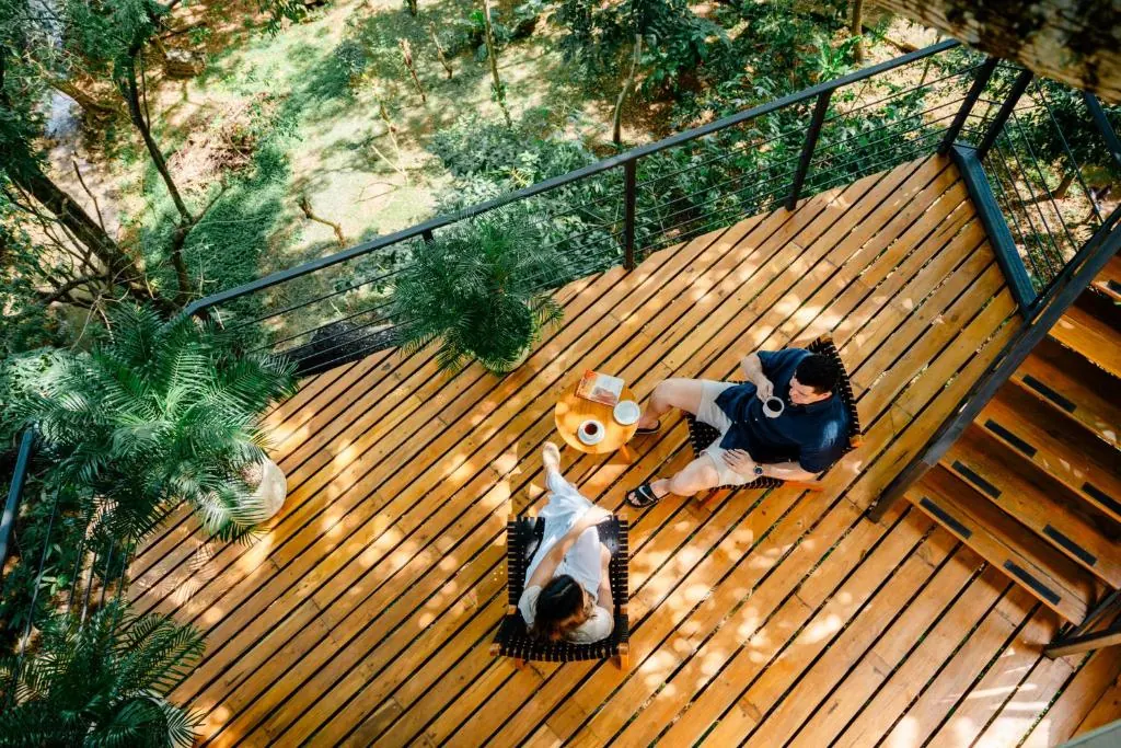 Two people sharing a slow morning tea ritual on a wooden treehouse deck, seen from above
